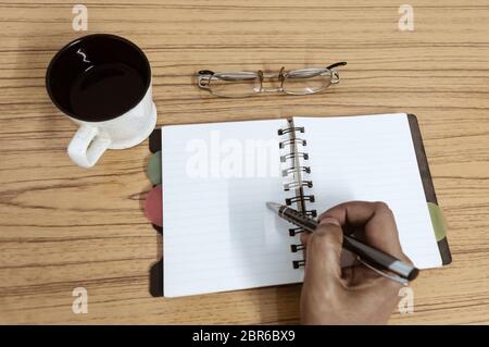 Imprenditore iscritto nel suo diario. Aprire il notebook con pagine vuote accanto alla tazza di caffè e occhiali sul tavolo di legno. Business ancora il concetto di vita WIT Foto Stock