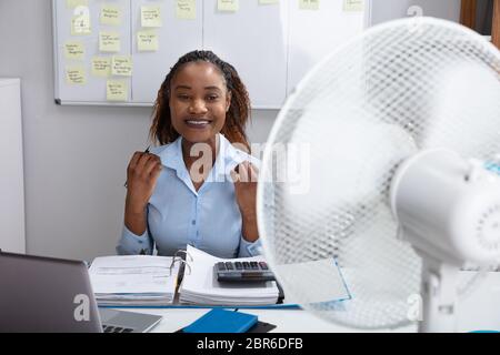 Giovane imprenditrice seduta in Office godendo brezza con ventola elettrica Foto Stock