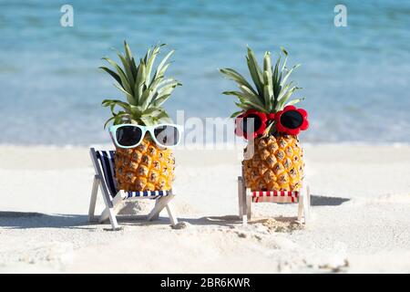 Due ananas con occhiali da sole sulle sedie a sdraio sulla sabbia in spiaggia Foto Stock