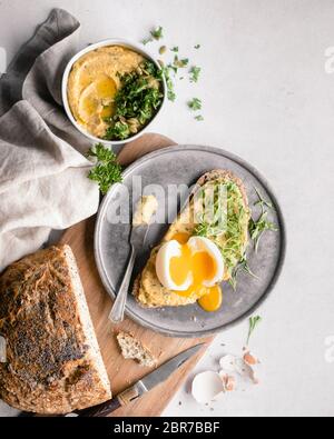 Una fetta di pane ricoperta di hummus, erbe e un uovo bollito morbido, circondato da filone tagliato, gusci di uova, un tovagliolo e una piccola ciotola di hummus su un wh Foto Stock
