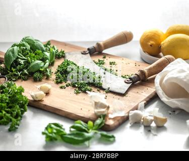 Un coltello con due manici poggiati su un mucchio di erbe verdi macinate su un tagliere di legno, circondato da limoni, spicchi d'aglio e un tovagliolo bianco Foto Stock