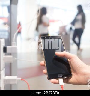 Mani femminili holding e utilizza lo smartphone durante la ricarica è in un luogo pubblico tramite spina elettrica e un cavo di ricarica. Foto Stock