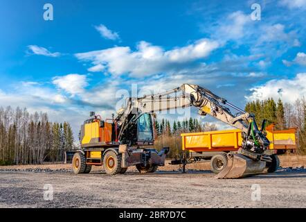 Bellissimo escavatore giallo grande con ruote grandi si trova vicino al carrello di servizio giallo su una nuova strada non ancora pronta. Serata soleggiata con nuvole blu, primavera Foto Stock