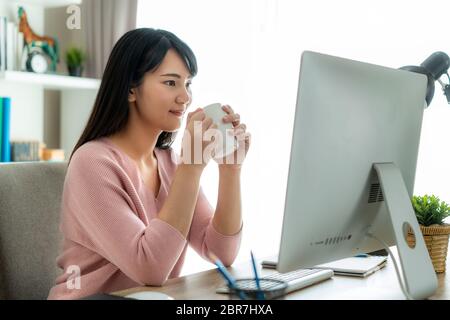 Asian bella giovane donna lavorare da casa lavorare sul computer e bere caffè mentre si lavora in soggiorno a casa. Stile di vita di distanza sociale i Foto Stock