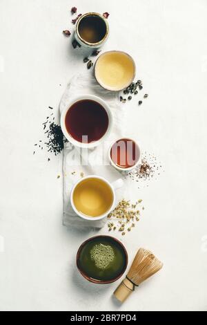 Tazze di tè con tè aromatico secco in ciotole su sfondo bianco vintage. Tè alle rose, tè verde, tè nero, tè rooibos, tè alla camomilla e tè alla matcha. Foto Stock