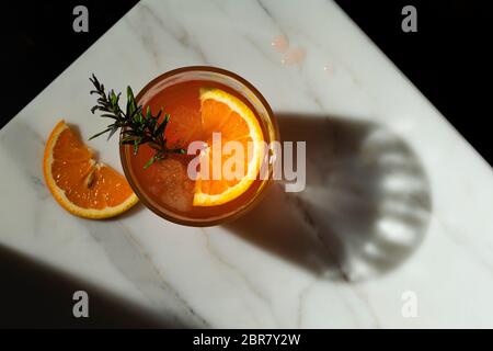 Cocktail Negroni con una fetta di arancia e una decorazione di sprig al rosmarino fumante. La vista è dall'alto, guardando verso il basso sull'angolo di un piano d'appoggio in marmo w Foto Stock