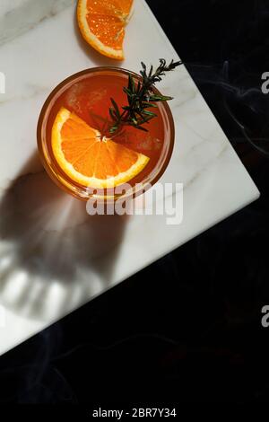 Cocktail Negroni con una fetta di arancia e una decorazione di sprig al rosmarino fumante. La vista è dall'alto e si affaccia su un piano portapaziente in marmo con una sezione di Foto Stock