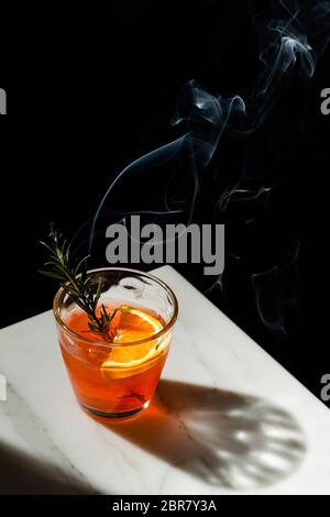 Cocktail Negroni con una fetta di arancia e una decorazione di sprig al rosmarino fumante. La vista presenta l'angolo di un piano portapaziente in marmo con l'ombra di Foto Stock