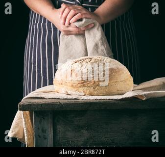 La donna in un grembiule detiene un lino grigio igienico accanto al forno pane tondo su una tavola di legno, sfondo scuro Foto Stock