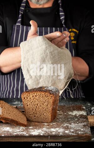 Chef in nero uniforme fette di pane di segale fatto su un marrone di legno cosparso di farina bianca e pulisce le mani con una corteccia grigia igienico, nero bac Foto Stock