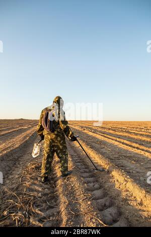 L'uomo in camuffamento con un metal detector e pala cammina attraverso il campo alla ricerca di valori antichi, nella sera d'estate. Foto Stock