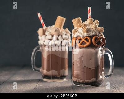 Vista ravvicinata dei due freakshake a mason jar sul tavolo marrone. Freaked frullato al cioccolato, pretzel, marshmallow, popcorn e waffles. Cibo alla moda Foto Stock