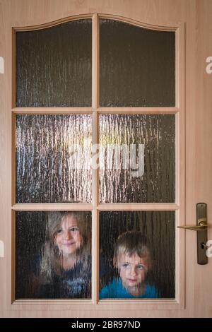 Ritratto di un grazioso piccolo ragazzo caucasico e bambina che si nasconde dietro una porta con finestre di vetro mentre si gioca a nascondino Foto Stock