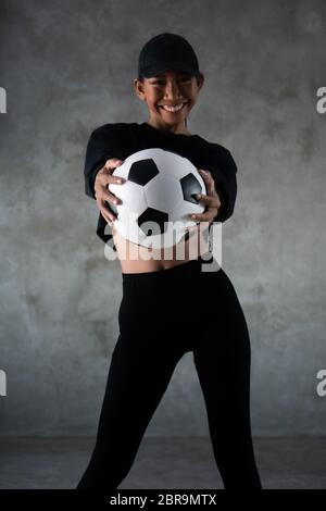 Ritratto di bella giovane donna sorridente in abiti sportivi tenendo la palla di calcio isolato su sfondo concreto Foto Stock