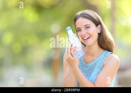 Donna felice che mostra una bottiglia d'acqua in plastica in un parco con uno sfondo verde Foto Stock