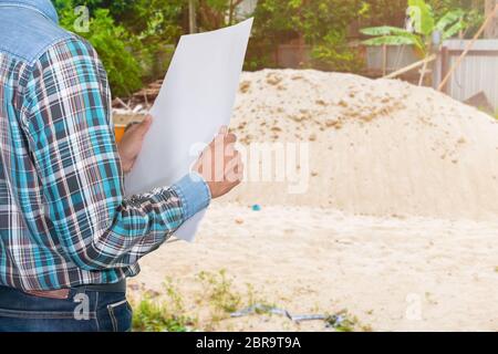 un tecnico di mano che tiene i progetti arrotolati e indossa plastica bianca per casco di sicurezza nel sito di ispezione Foto Stock