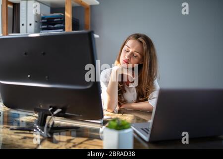 Stanco giovane imprenditrice lavorando in ufficio con computer e laptop sulla scrivania in ufficio Foto Stock