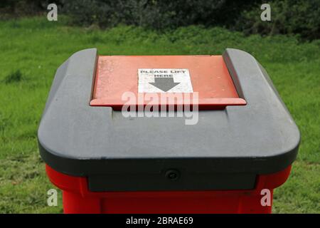 Red Dog litter bin in close up visto dalla parte anteriore con adesivo consultivo e coperchio nero con rosso incernierato scivolo. Sullo sfondo di un parco con prato. Foto Stock