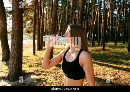 Vista laterale della giovane donna attraente bevendo acqua dalla bottiglia dopo esercizio nel parco, all'esterno. Vestito in abbigliamento sportivo. Foto Stock