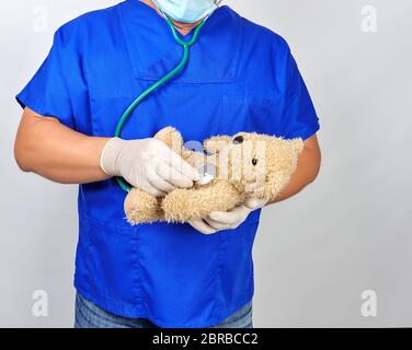 Dottore in uniforme blu e bianco guanti in lattice tenendo un marrone Teddy bear, pediatra ascolta con un giocattolo stetoscopio Foto Stock
