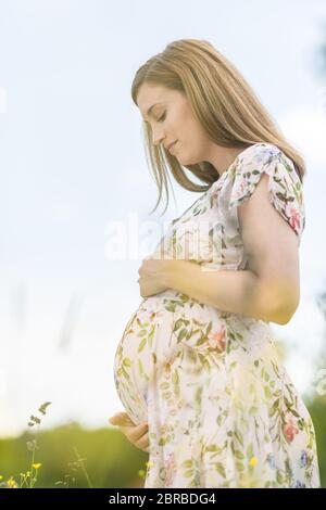 Ritratto di bella donna incinta in bianco abiti estivi rilassante in prato pieno di giallo flovers fioritura. Concetto di sana maternità cura. Foto Stock