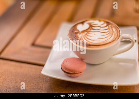 Primo piano di cappuccino latte caldo con bella latte art in una tazza di ceramica bianca su uno sfondo di tavola di legno e biscotti macaron Foto Stock