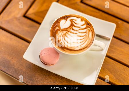 Primo piano di cappuccino latte caldo con bella latte art in una tazza di ceramica bianca su uno sfondo di tavola di legno e biscotti macaron Foto Stock