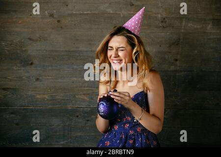 Felice ragazza prorompente di ridere mentre cercando di far esplodere un palloncino, indossare un abito carino, festosa hat. Foto Stock