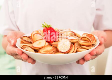 Bambino che tiene il recipiente bianco con i mini pancake sani alla moda cereali e fragole. Nessuna immagine del volto, concetto di famiglia. Sana colazione vegana senza Foto Stock