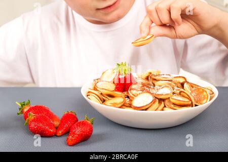 Bambino ragazzo che mangia minifittelle senza glutine cereali con fragole da ciotola bianca. Nessuna immagine del volto, concetto di famiglia. Sana e trendy colazione vegan Foto Stock