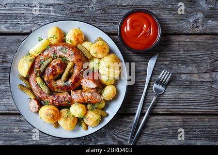 Cena con salsiccia di patate e cetriolini nuovi, cosparsa di aneto fresco su un piatto con posate e ketchup su un tavolo di legno, vista dall'alto, primo piano Foto Stock