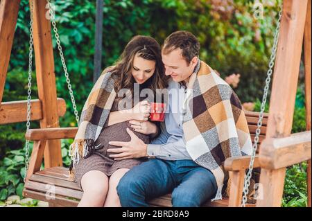 Una giovane donna incinta e suo marito. Una famiglia felice seduta su un altalena, tenendo il ventre. Donna incinta rilassarsi nel parco. Foto Stock