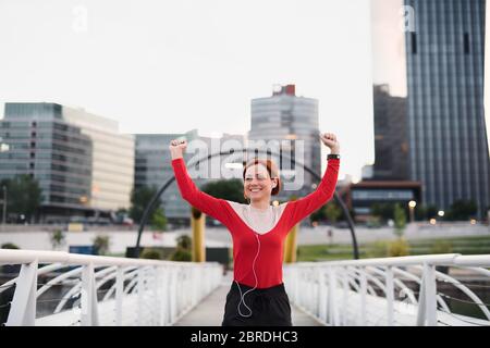 Vista frontale della giovane donna runner con auricolari in città, riposante. Foto Stock