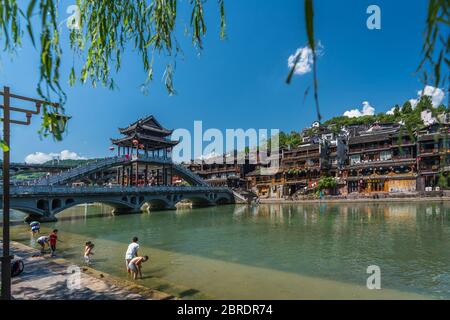 Feng Huang, Cina - Agosto 2019 : Bambini e residenti locali si lavano, spruzzano e si divertono in acqua accanto al ponte ad arco panoramico sopra Tuoji Foto Stock