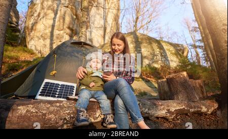 Donna e bambino piccolo sono seduti su un ceppo vicino tenda turistica e pannello solare ai piedi di una scogliera nella foresta in una giornata di sole. La mamma mostra al bambino qualcosa su un telefono cellulare Foto Stock