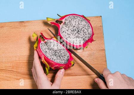 Pitaya tagliato in due metà. Primo piano di mani femminili che tagliano frutta estiva del drago. Pitaya su una tavola di legno con sfondo blu Foto Stock