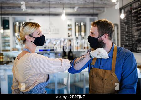 I proprietari di una caffetteria con maschere facciali che battono il gomito, aprono dopo la quarantena. Foto Stock