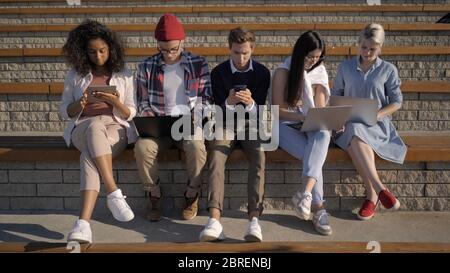 Studenti multietnici seduti in fila assorbiti da telefoni, compu Foto Stock