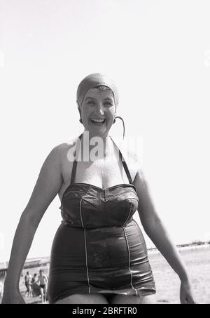 1961, storica, una donna matura, piuttosto portly in un costume da bagno bagnato e cappello di gomma sorridente, avendo appena fatto una nuotata in mare, Inghilterra, Regno Unito. Foto Stock