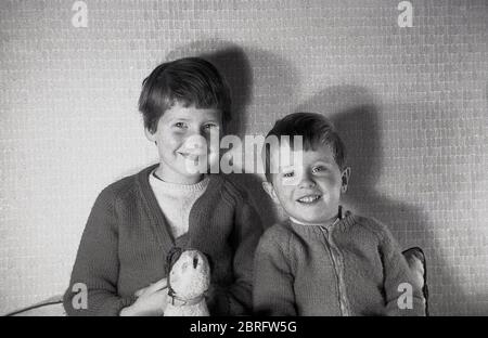 Anni '60, storico, due voi bambini, un bambino e la sua sorella leggermente più grande sorridendo come si pongono insieme per la foto in interni, con la giovane ragazza che tiene il suo giocattolo morbido, in una foto che riassume la semplice e felice innocenza dell'infanzia trovato in questa epoca, Inghilterra, Regno Unito. Foto Stock