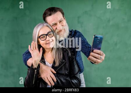 Ritratto di una coppia anziana attraente, in piedi a casa davanti alla parete verde e parlare con i loro amici tramite chat video telefonica. Uomo bearded Foto Stock