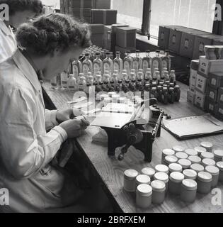 Anni '50, lavoratrice storica, femminile in un cappotto bianco seduto su un banco di lavoro di legno etichettare piccole bottiglie di medicinali usando una piccola macchina manuale, Inghilterra, Regno Unito. Foto Stock