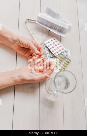 Mani senior con pillole e farmaci sul tavolo, bicchiere d'acqua. Le mani rughe della vecchia donna che tiene compresse colorate, occhiali e prescrizione di legno indietro Foto Stock