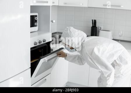 L'uomo in tuta protettiva bianca e in faccia maschera medica cuoce nel forno nella cucina bianca a casa durante il coronavirus. Rimani a casa. Goditi la cucina Foto Stock