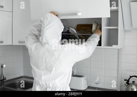 L'uomo in tuta protettiva bianca e in faccia maschera medica cuoce nella cucina bianca a casa durante il coronavirus. Rimani a casa. Goditi la cucina a casa Foto Stock