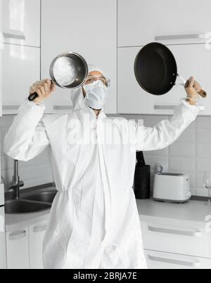 L'uomo in tuta protettiva bianca e in maschera medica di faccia tiene due padelle frittura nella cucina bianca a casa durante il coronavirus. Rimani a casa. Divergetevi Foto Stock