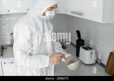 L'uomo in tuta protettiva bianca e in faccia maschera medica cuoce nella cucina bianca a casa durante il coronavirus. Rimani a casa. Goditi la cucina a casa Foto Stock