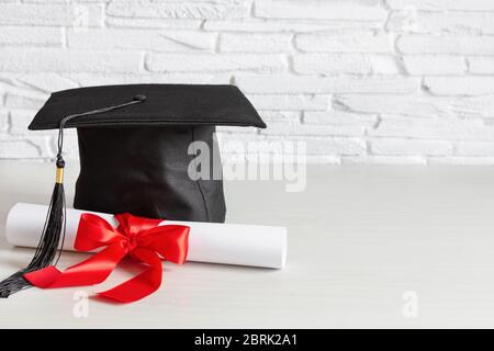 Cappellino accademico e diploma di laurea scroll e legato con nastro rosso su tavolo di legno bianco. Foto Stock