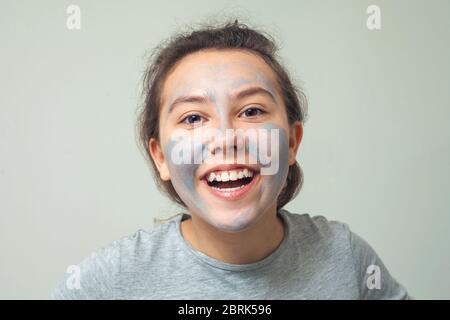 Cute e felice ragazza striscio faccia con argilla cosmetica o fango e sorrisi. Maschera cosmetica, scrub viso. Il concetto di salute e bellezza. Foto Stock