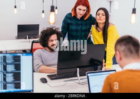 Due giovani donne d'affari accanto a un collega maschile mentre spiega le nuove attività online su un computer in un ambiente di ufficio confortevole. Foto Stock